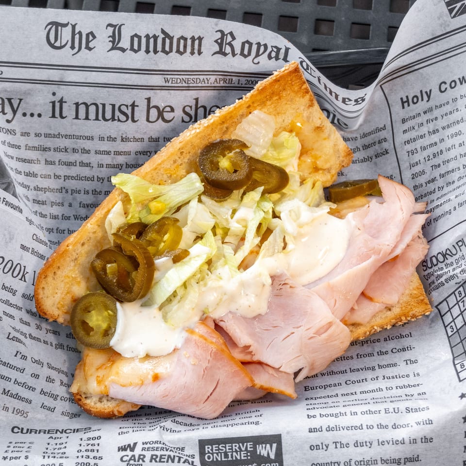 A turkey sandwich with lettuce, jalapeños, and white sauce on toasted bread, served on newspaper.