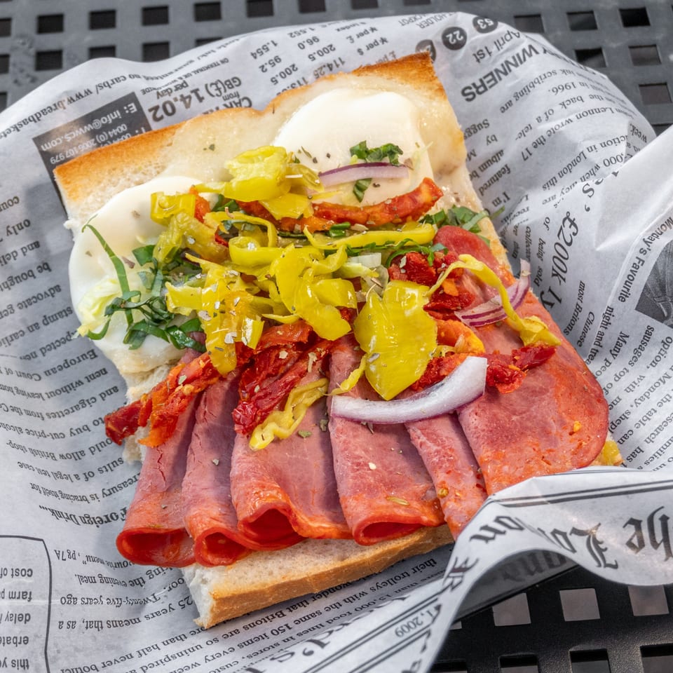 A close-up of a deli sandwich with sliced meats, cheese, peppers, and herbs, wrapped in newspaper.