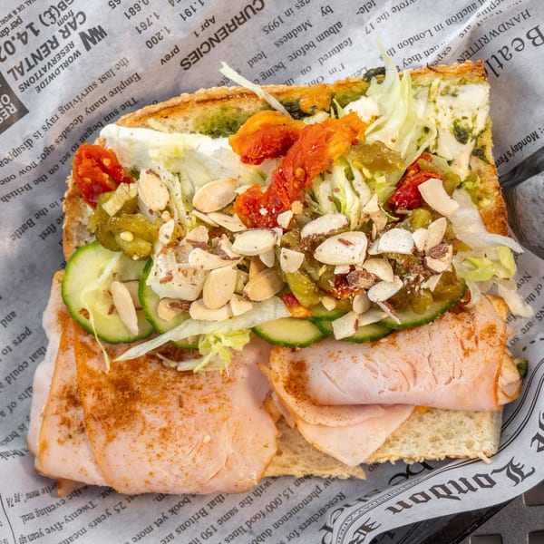A turkey sandwich with pesto, sun-dried tomatoes, cucumbers, and almonds on newspaper-patterned paper.
