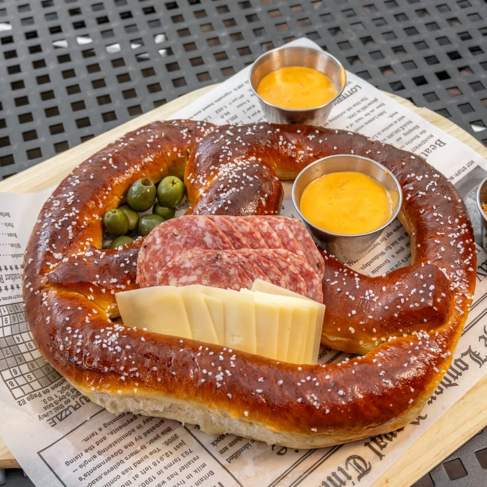 A large pretzel with cheese, salami, olives, and two dipping sauces on a newspaper-lined tray.