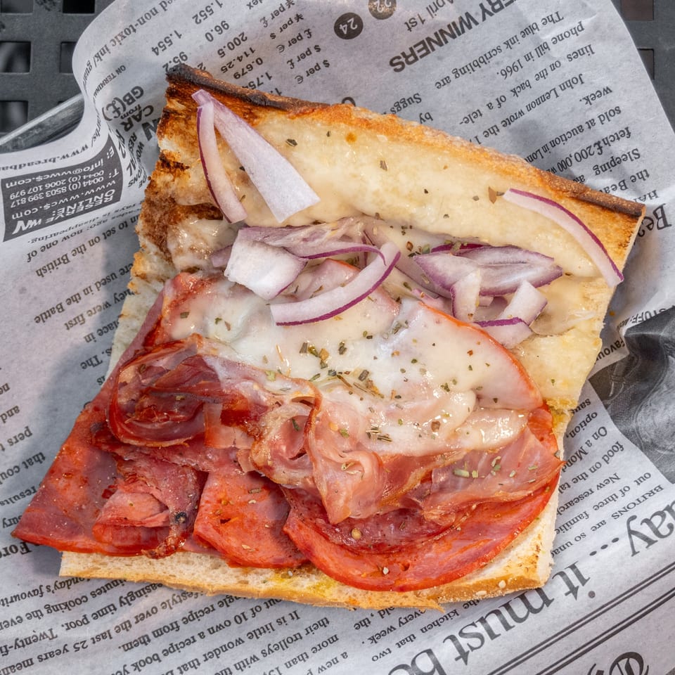 A toasted sandwich with melted cheese, red onions, and cured meats, served on newspaper.
